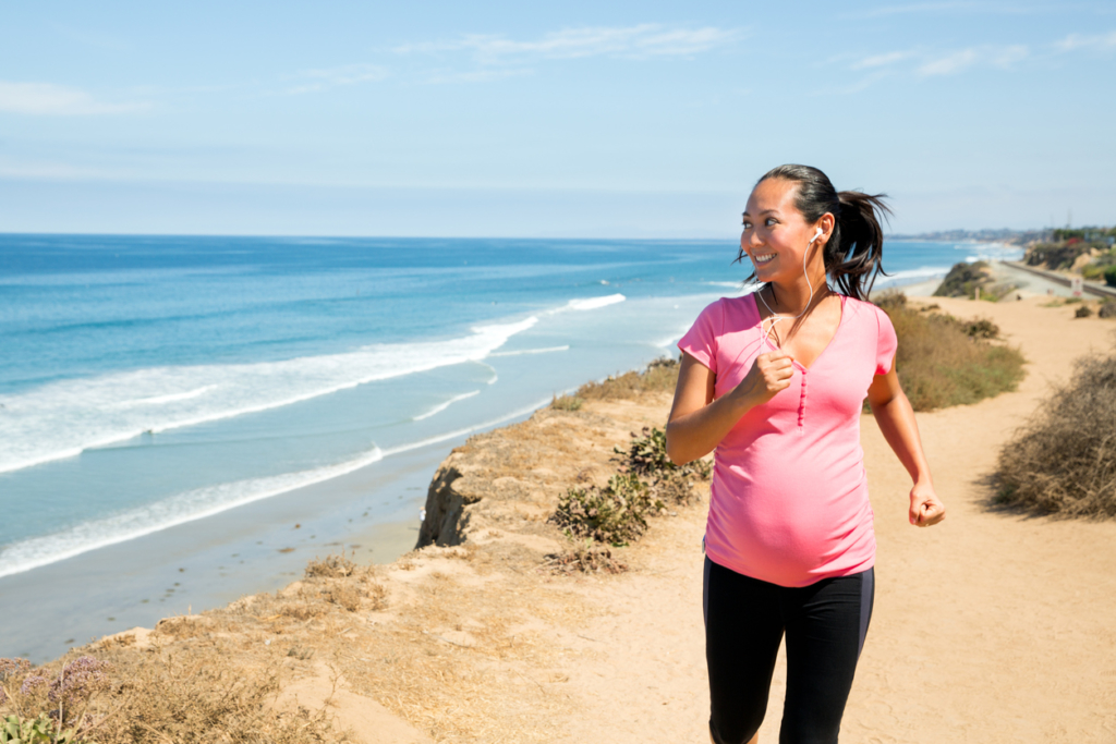A beautiful pregnant woman running on the nature trail overlooking the ocean in San Diego, California.  She is in her third trimester (7 months) and wearing a pink maternity sports top and leggings. She's been a runner and continues to jog regularly to stay fit and strong during pregnancy. Being physically active is important for a healthy pregnancy. She loves looking at the ocean as she runs.
