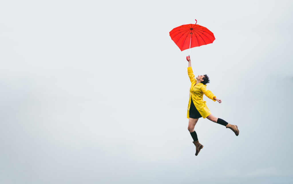 Surreal moment of an young caucasian woman flying in the middle of the clouds hanging on her umbrella.